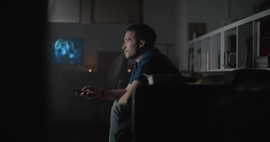 Tired man sitting alone on the couch in the living room using a remote control and watching tv