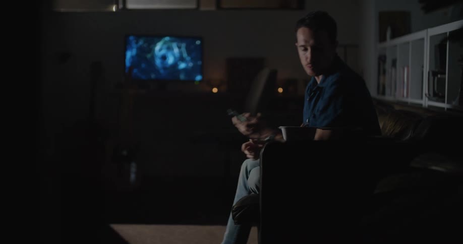A man texting on his phone while eating in his living room at night