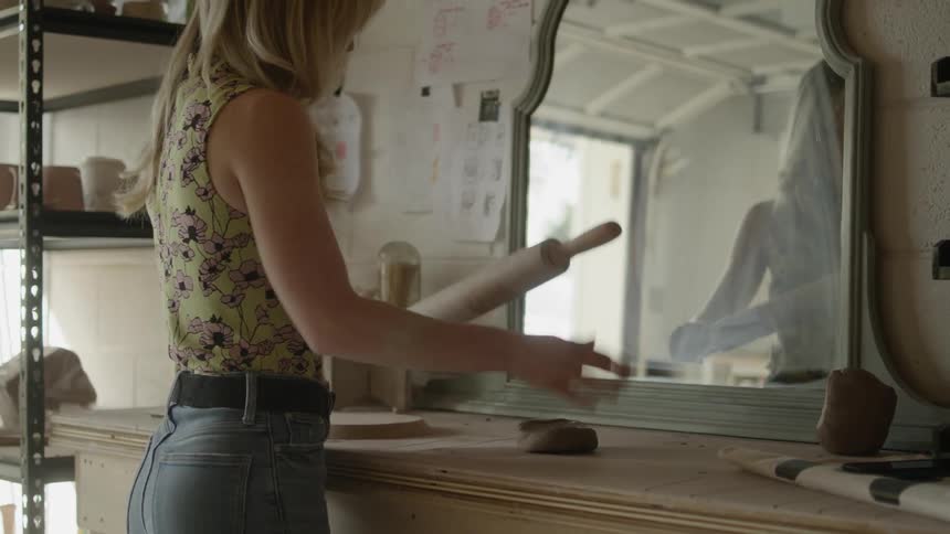 A woman with blonde hair using a rolling pin to flatten a piece of sculpting clay