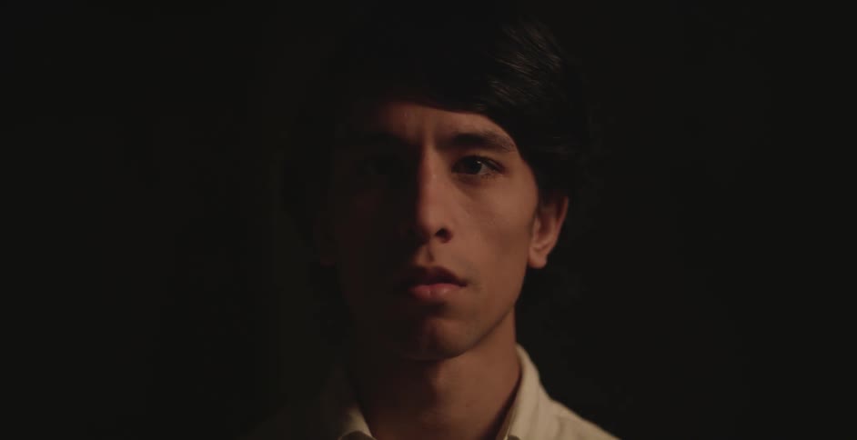 A man is facing forward in a dark area half of his face is in shadow