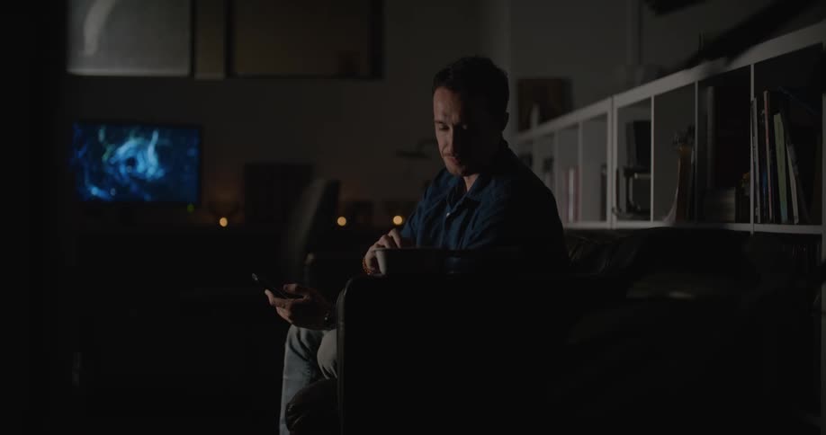 A man sits on a couch in front of his television leans forward while looking at his phone