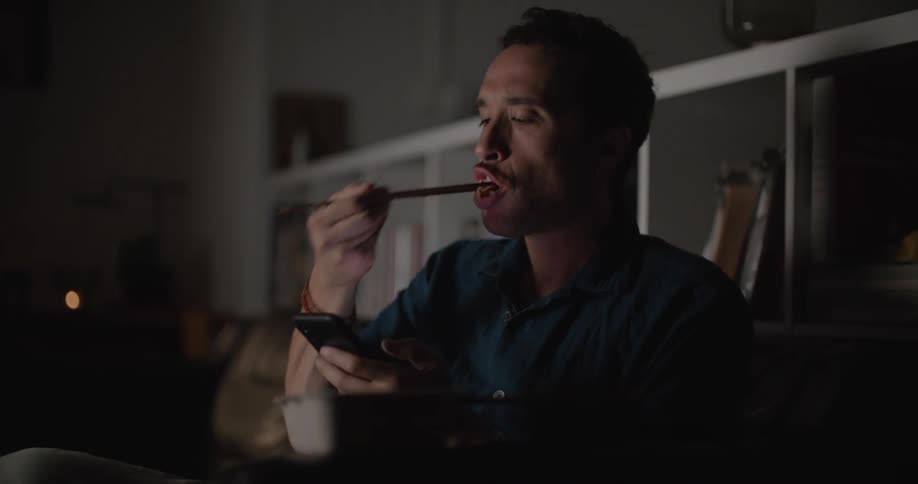 Man with mustache and dark shirt eats food with chopsticks using smartphone in living room at home