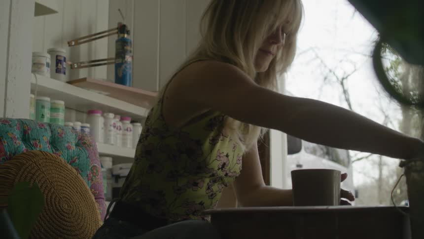 A woman with long blonde hair carefully shapes a block of clay as she sits indoors