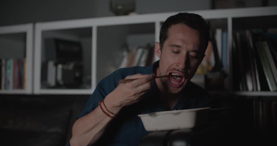 A man sits on a couch in front of his television while eating takeout with chopsticks and looking at his phone