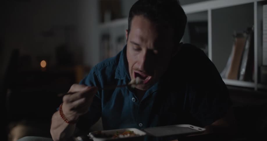 A man sits on a couch in front of his television while eating and looking down at his phone