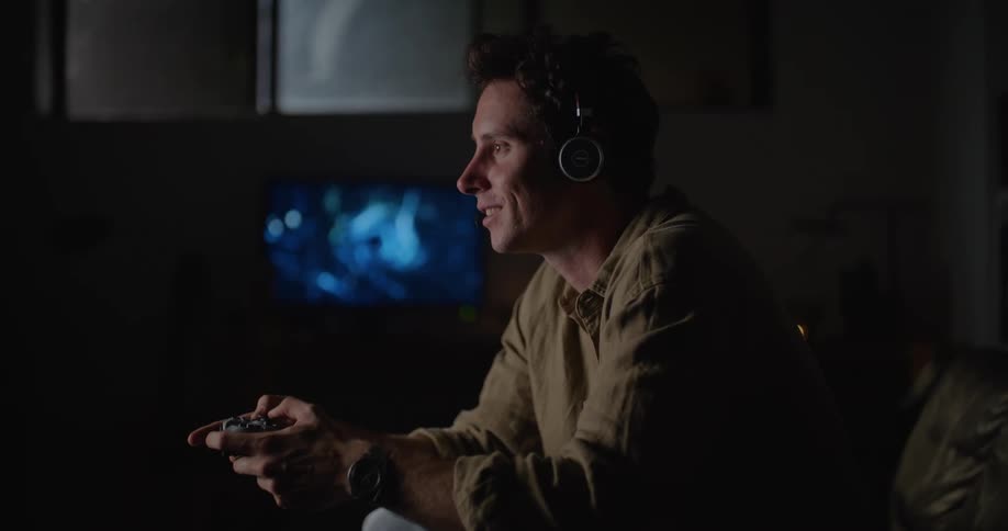 A man talks into his headset while playing a video game in the dark living room of a house at night