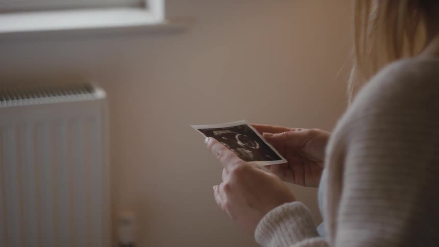 Pregnant woman with brown hair and gray sweater looks at ultrasound baby photograph