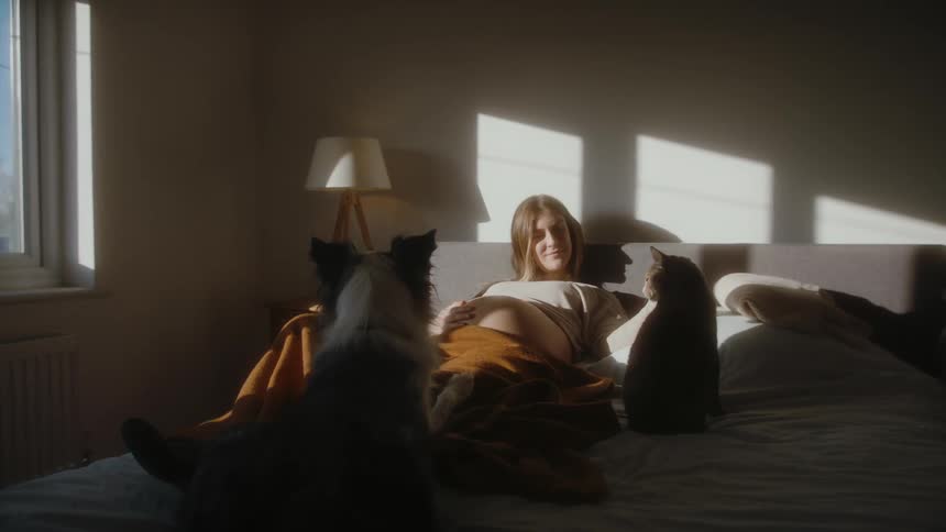 A pregnant woman pets a cat while resting in bed with a cat and dog
