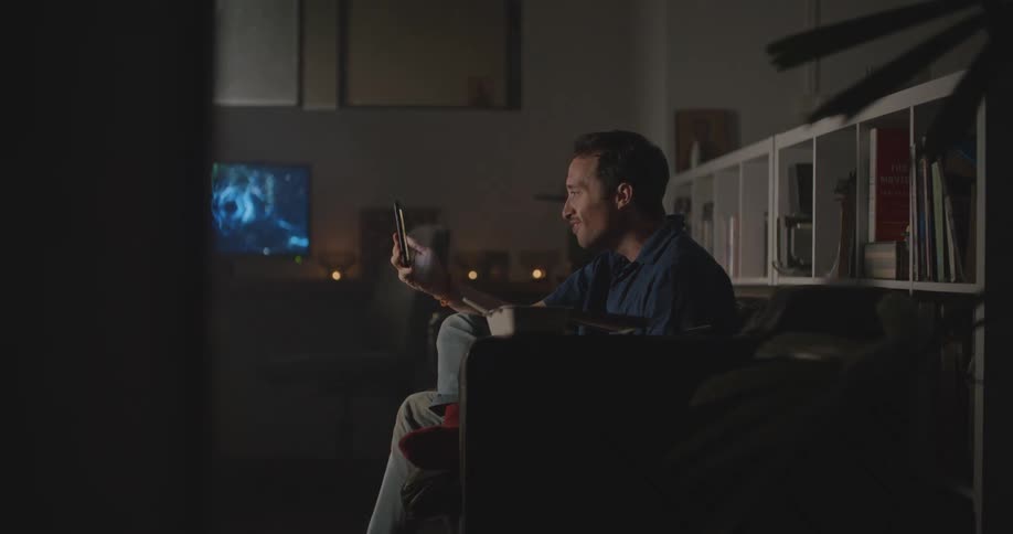 Man with blue shirt and jeans sits on couch eating food and making video call on smartphone in living room at home