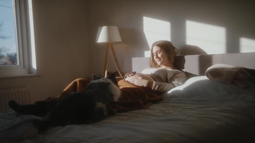 A pregnant woman strokes her belly while resting in bed with a dog