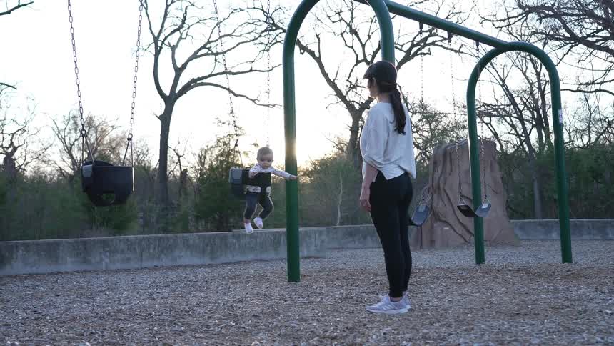 Mother with ponytail white shirt and black jeans swings baby in playground at dusk