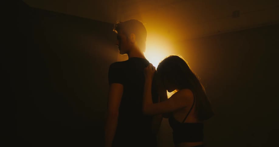 Woman with black dress rests her head against mans back wearing black t shirt in spotlight