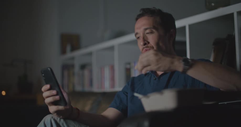 A man eats while participating in a video chat on his smartphone on the sofa of a dark living room