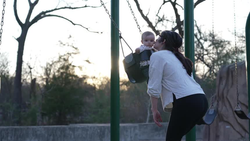 A baby swings in a swing and as she swings forwards her mother bends down and kisses her face