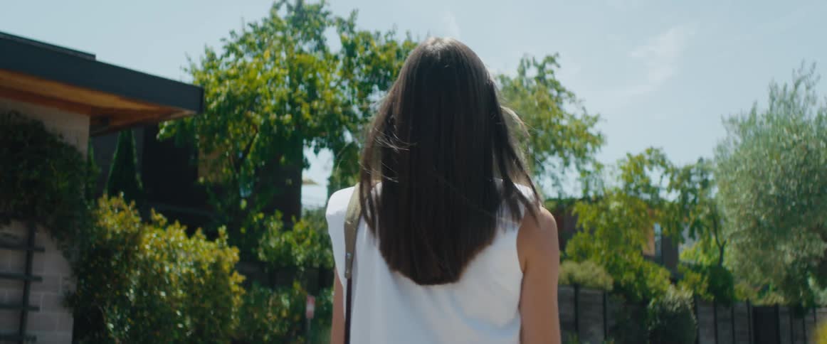 A woman walks outdoors beside a small building on a sunny day