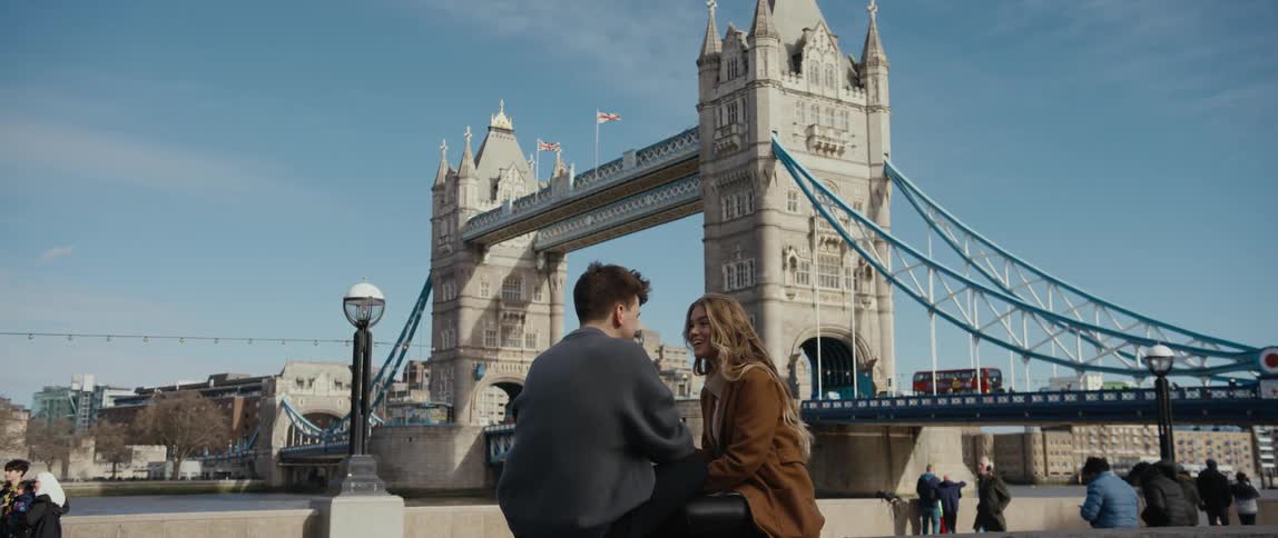A man and woman sit and talk and hold hands by a river as people stand and look at the river and walk past