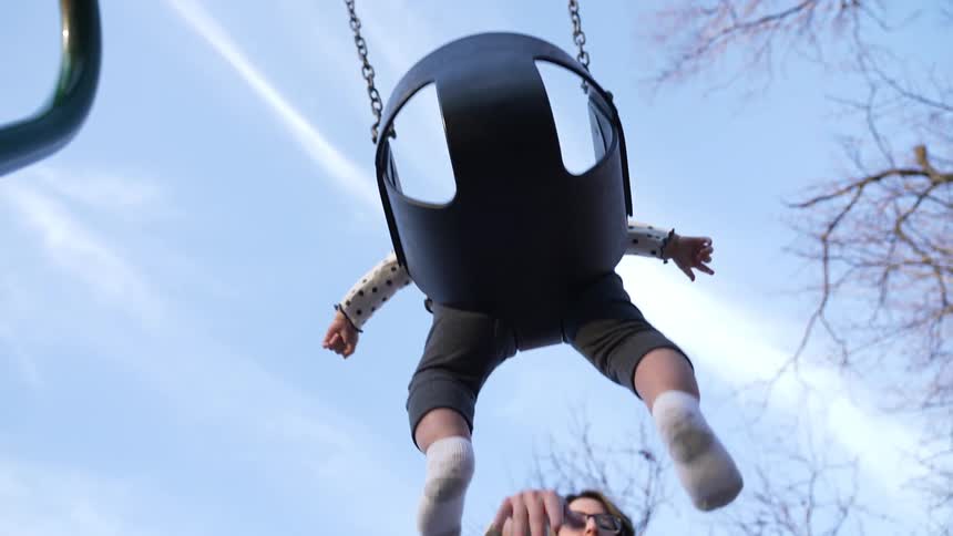 Mother pushes toddler in swing under leafless trees