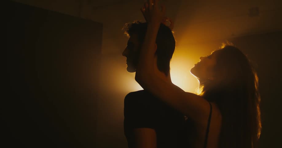 Man standing in the middle of an empty room lightened by an orange light while a woman is performing a dance move behind him