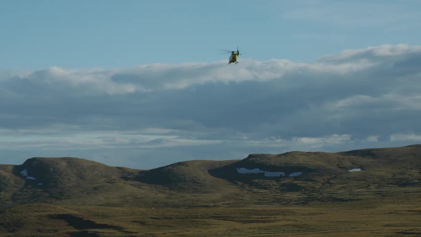 A helicopter flies over a hilly countryside on a cloudy day