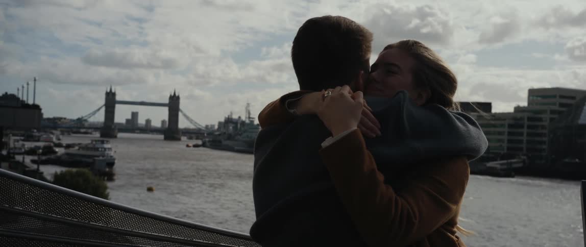 Man and woman embrace and hug as they stand near a river under a cloudy blue sky