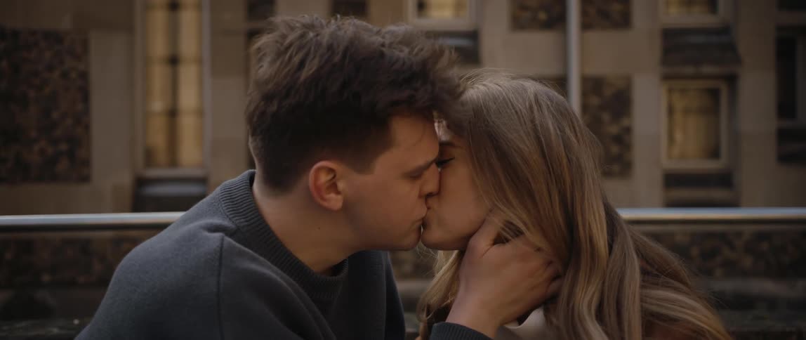 Couple gazes into each others eyes and kiss sitting near rail with building in background