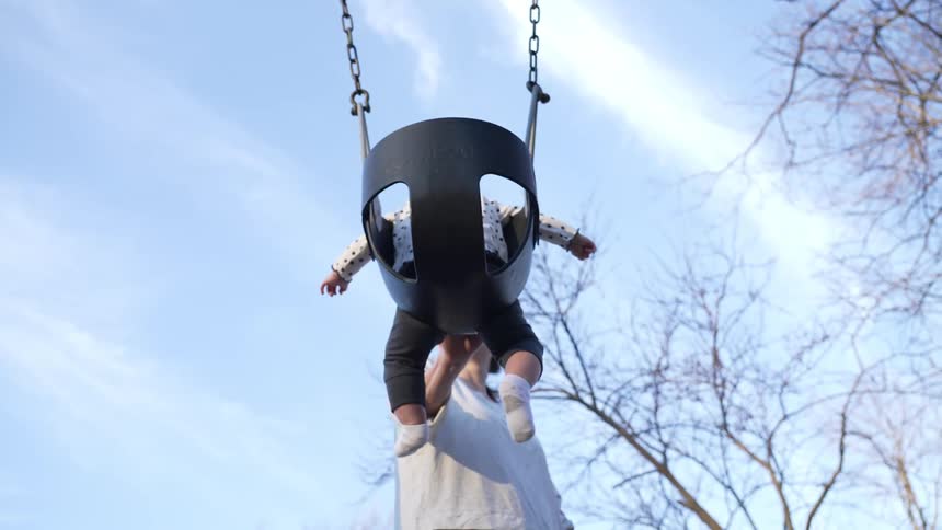 Mother pushing the fullbucket swing seat her baby is sitting on at an outdoor playground