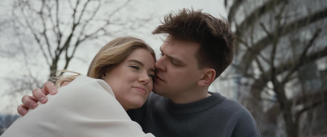 Woman hugs a man as he kisses her face while they sit near the bare trees on a breezy day