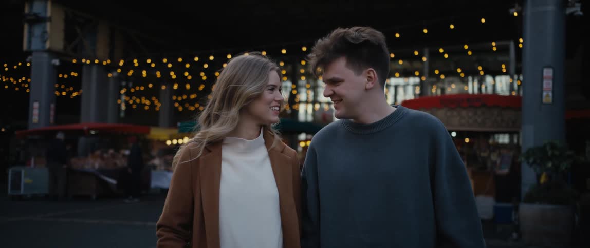 Man with gray sweater and blonde woman with white shirt and brown overcoat holding hands lovingly look into each other in front of shopping mall