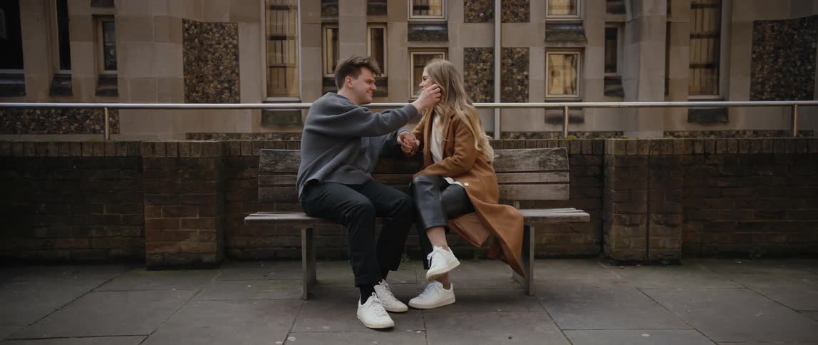 Man with gray sweater black pants white shoes and blonde woman wearing brown overcoat pants and white shoes sit on bench and lovingly look into each others eyes