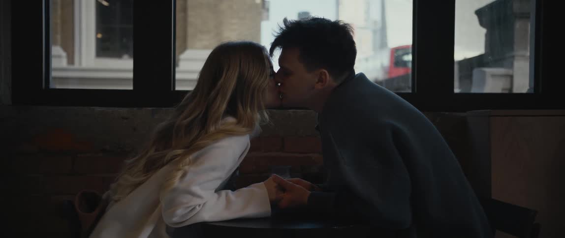 Blonde girl with white shirt and man with gray sweater sits at table lovingly looking into each others eyes and kiss each other at cafe