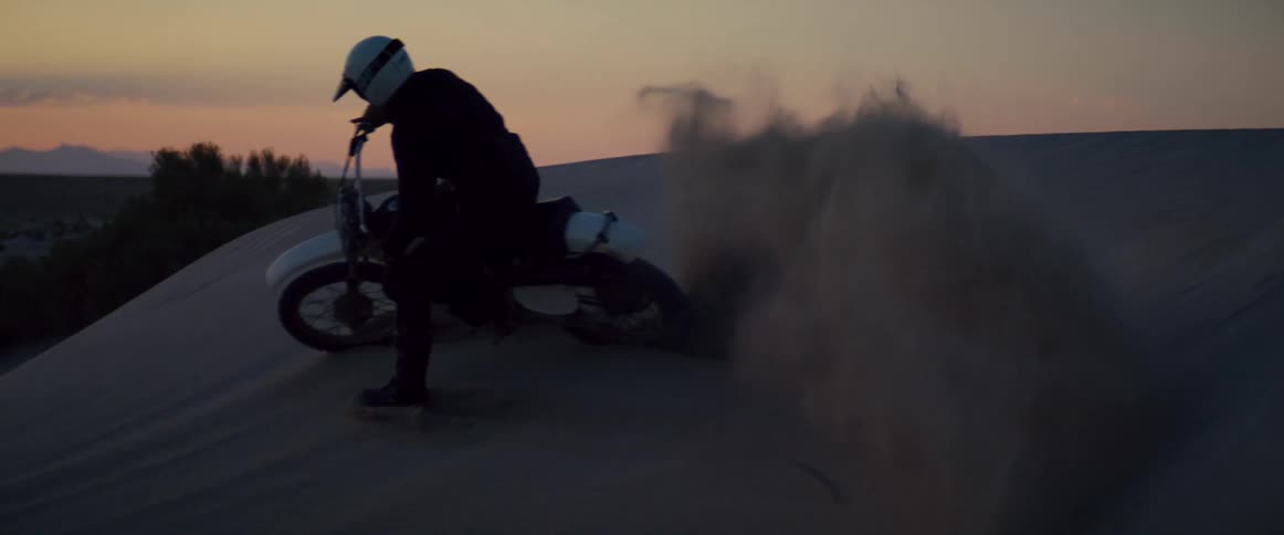 Person rides motorcycle up and down dune with scrub land and mountains in background under a red sky