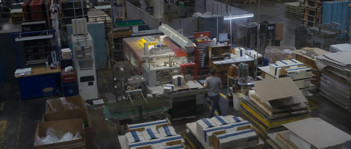 Woman stacks some bundles of paper on a table in a warehouse then walks around the table to check on something