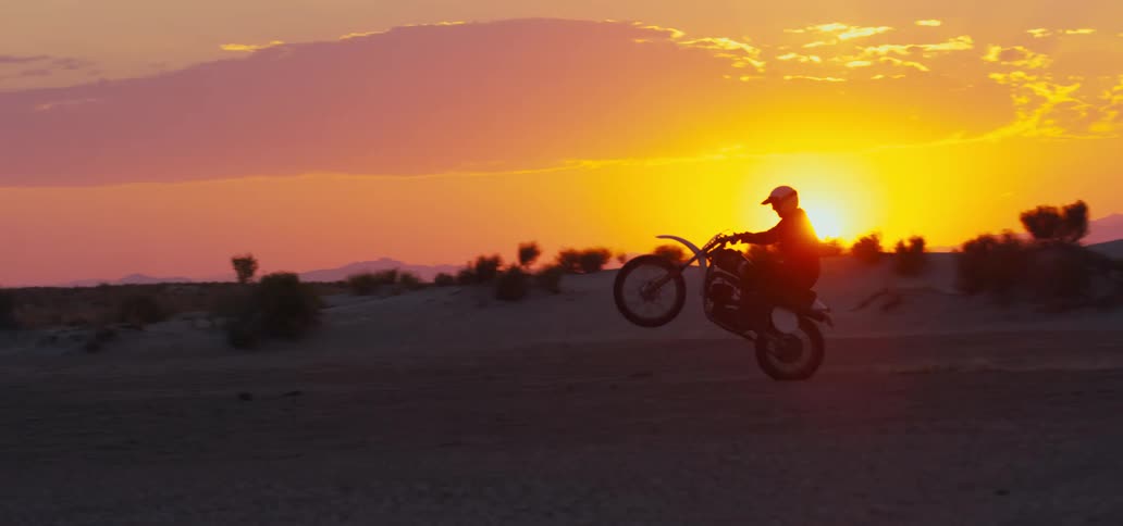 A motorcyclist does a wheelie while riding through the desert at sunset