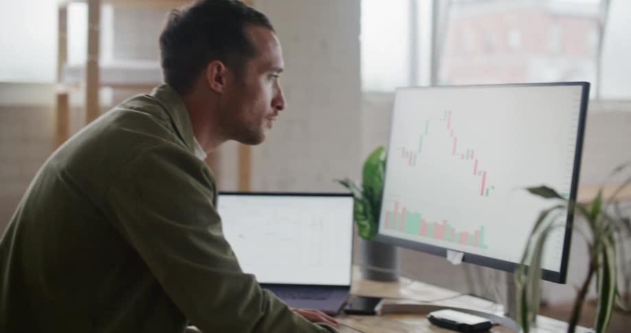 A man is sitting at a desk looking at a monitor with a graph on it and a laptop computer