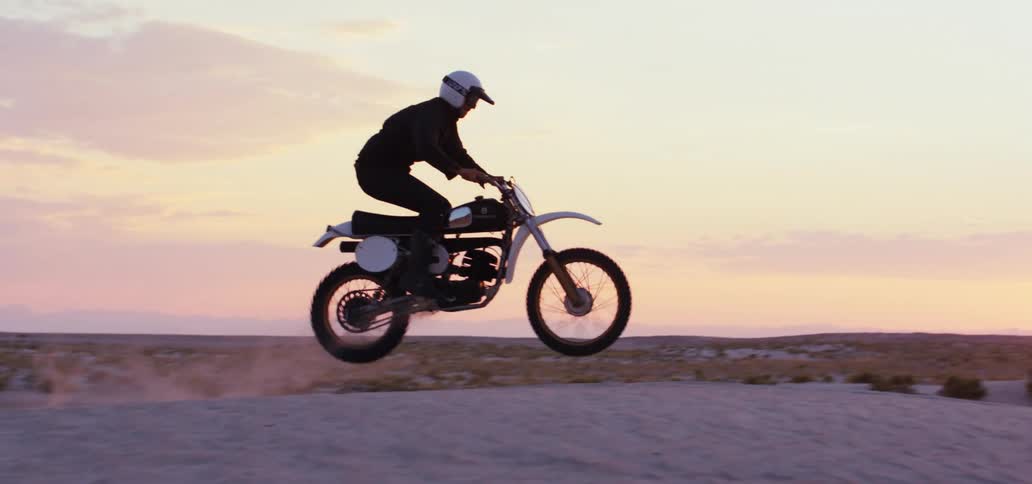 A motorcyclist briefly goes airborne while riding up a hill in an arid countryside at sunset