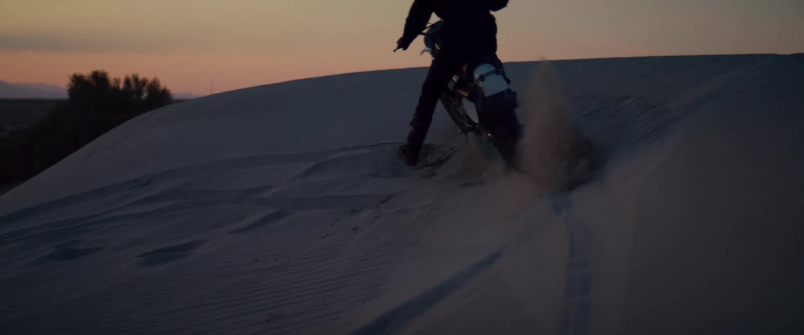 Person riding motorcycle rides up and down side of dune spraying sand into the air under red sky