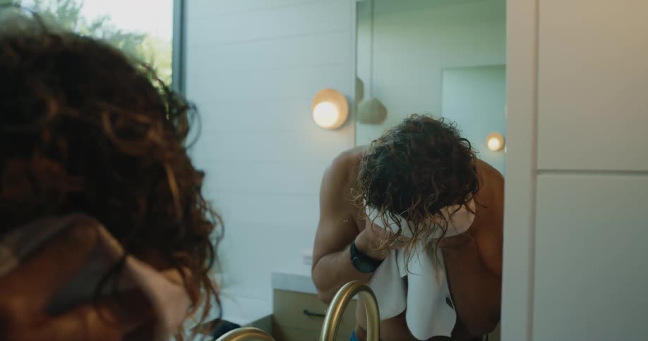 Shirtless man in the bathroom at home looking at himself in the mirror and drying his face with a hand towel