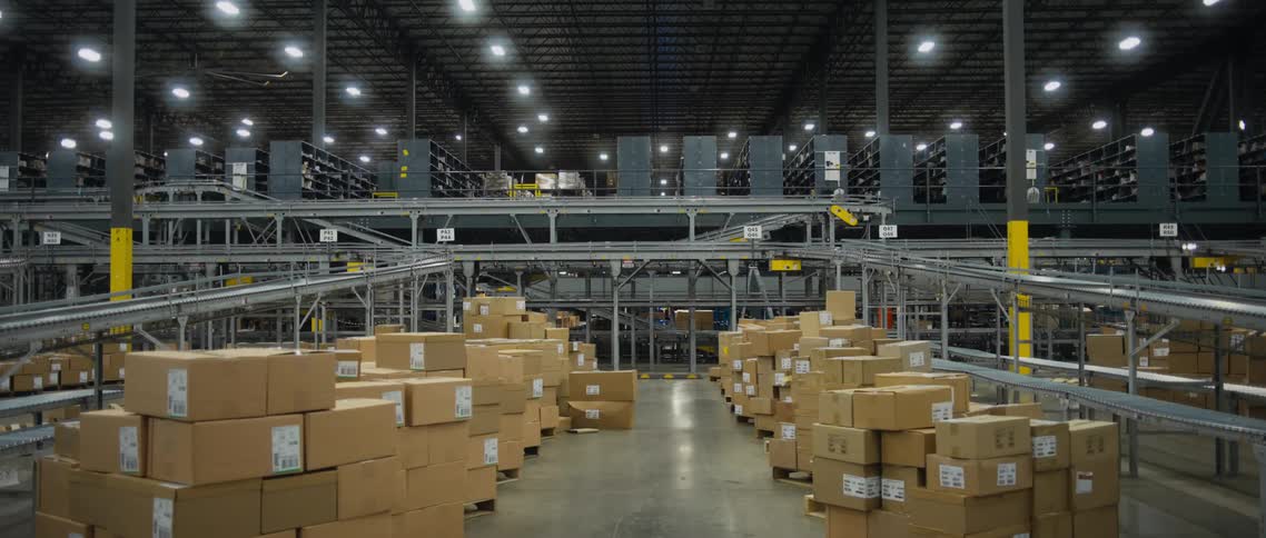 Stacks of boxes piled on a floor in a warehouse as workers move in the background