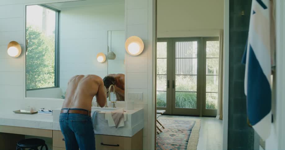 Shirtless man wearing jeans stands at the bathroom sink and washes his face