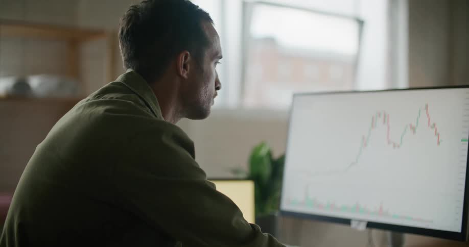 A man is sitting at a desk looking at a monitor with a graph on it and a laptop computer