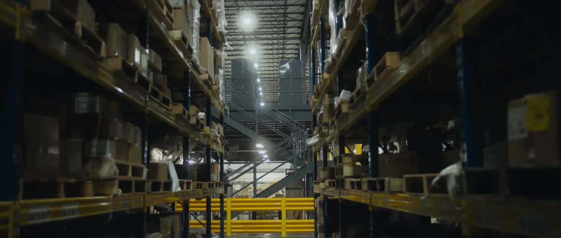 People walk past shelves in a warehouse lined with pallets