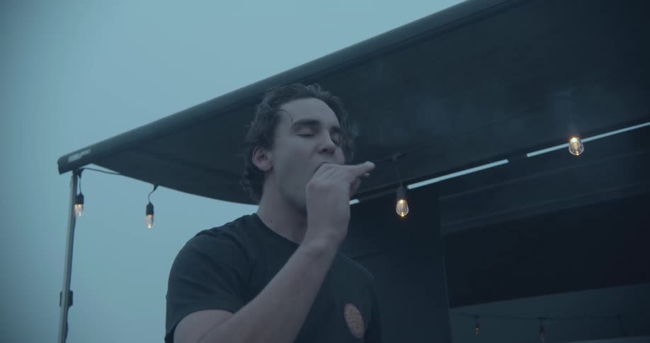 A man lights kindling in a campfire then places in his mouth and exhales smoke