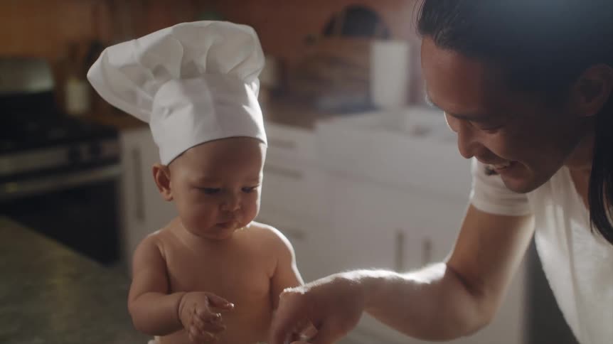 Father with long hair white t shirt and apron plays dough with baby son wearing chef hat and diaper sitting on kitchen island in the kitchen