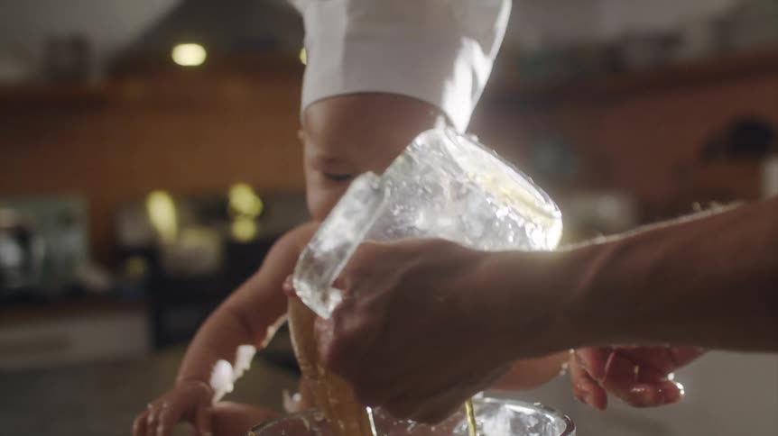 A man pours liquid from a measuring cup into a mixing bowl as his baby watches