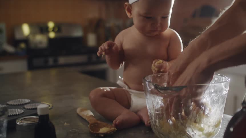 Man mixing food inside a glass bowl while his baby son is sitting right next to it at the kitchen counter eating something