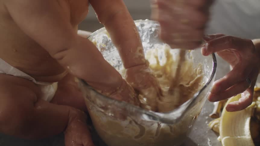 An adult uses a wooden mixing spoon to mix dough in a bowl as a baby tries to mix the dough with their hands