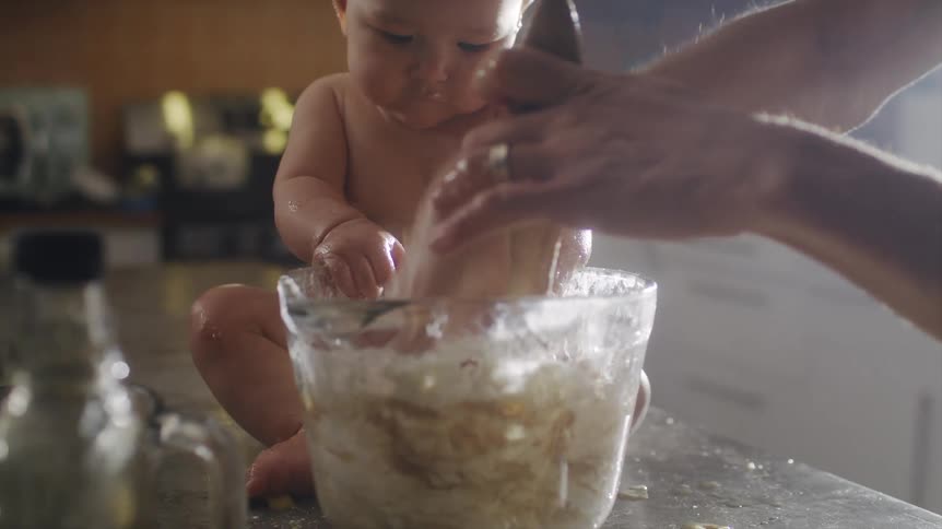 A baby sits on the kitchen counter and helps its dad mix batter in a bowl