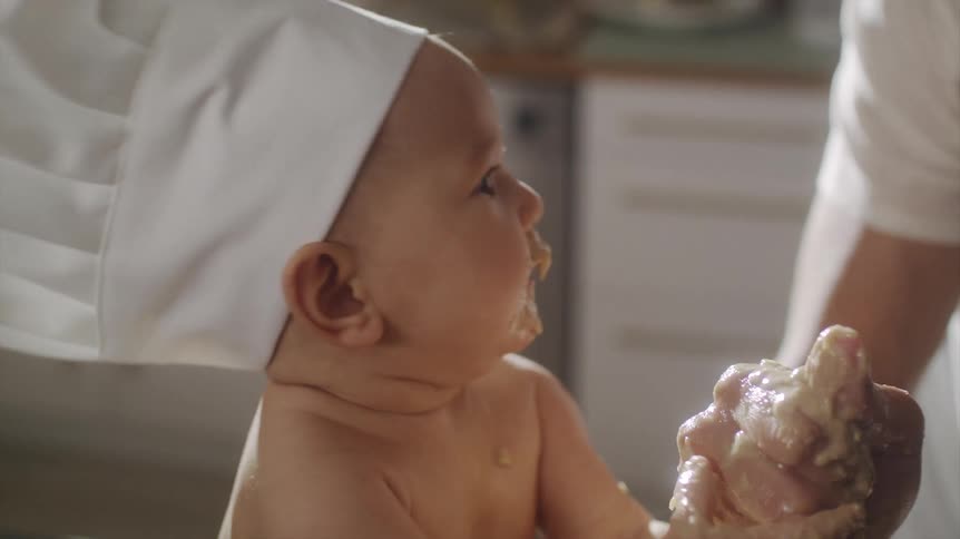 A man helps a baby in a chef hat to stir batter in a mixing bowl on a kitchen table