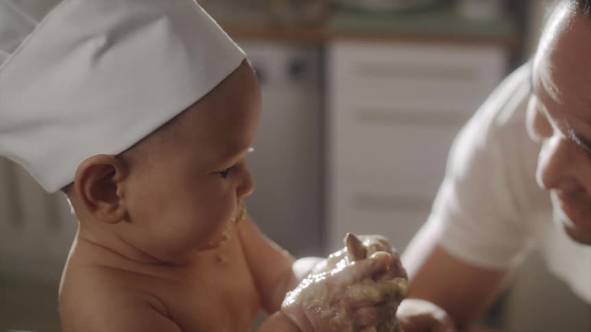 A man laughs as his baby eats batter off a mixing spoon