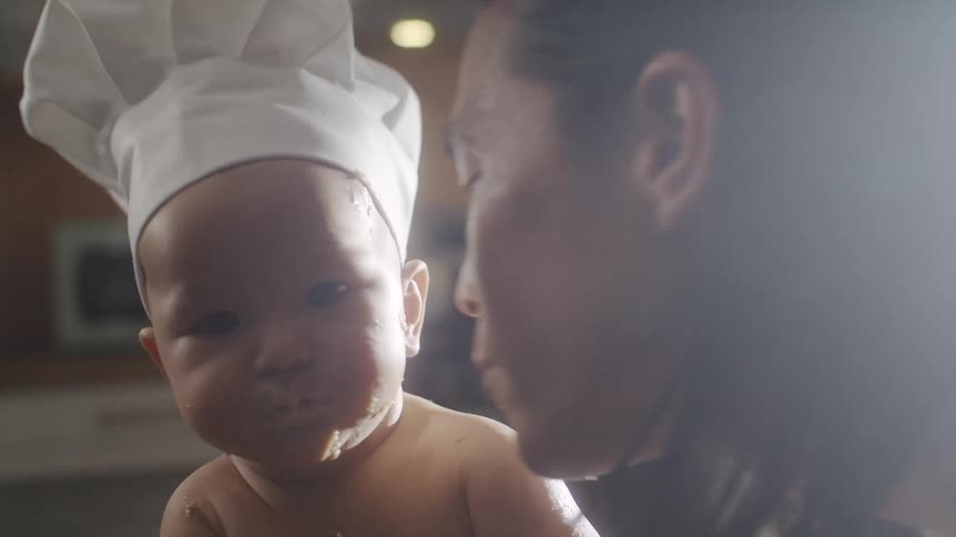 Baby covered in batter wearing a chef hat being kissed by his father
